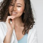Young woman with curly hair and eye patches smiling confidently in casual attire.